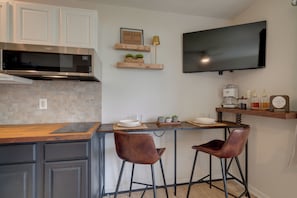 A modern kitchen with a microwave, two bar stools, a coffee station, and a flat-screen TV mounted on the wall.