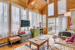 A wooden cabin interior with a TV, green cabinet, sofa, and a view through the glass door. - Cottage w/ Shared Beach, Great Views, Walk to Town (Schroon Lake)