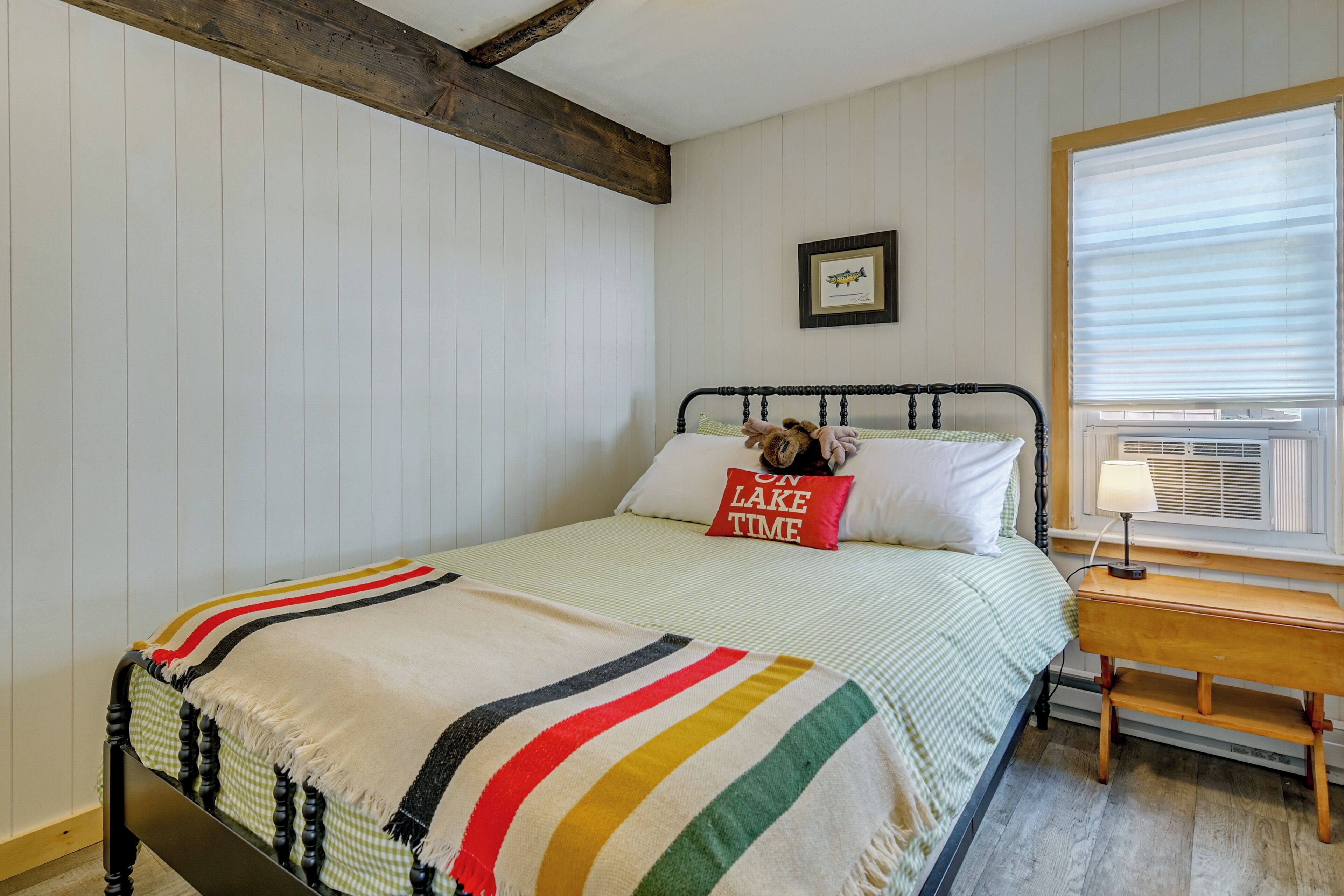 Een bed met een gestreept dekbed, een kussen met de tekst 'ON LAKE TIME', een klein nachtkastje met een lamp en een raam met luxaflex.