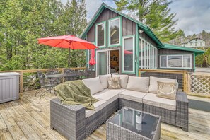 Una terraza de madera con mobiliario exterior, un paraguas rojo y una casa con ventanas amplias y techo verde.