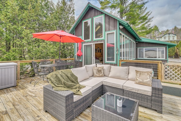 A wooden deck with outdoor furniture, a red umbrella, and a house with large windows and a green roof. - Cottage w/ Shared Beach, Great Views, Walk to Town (Schroon Lake)