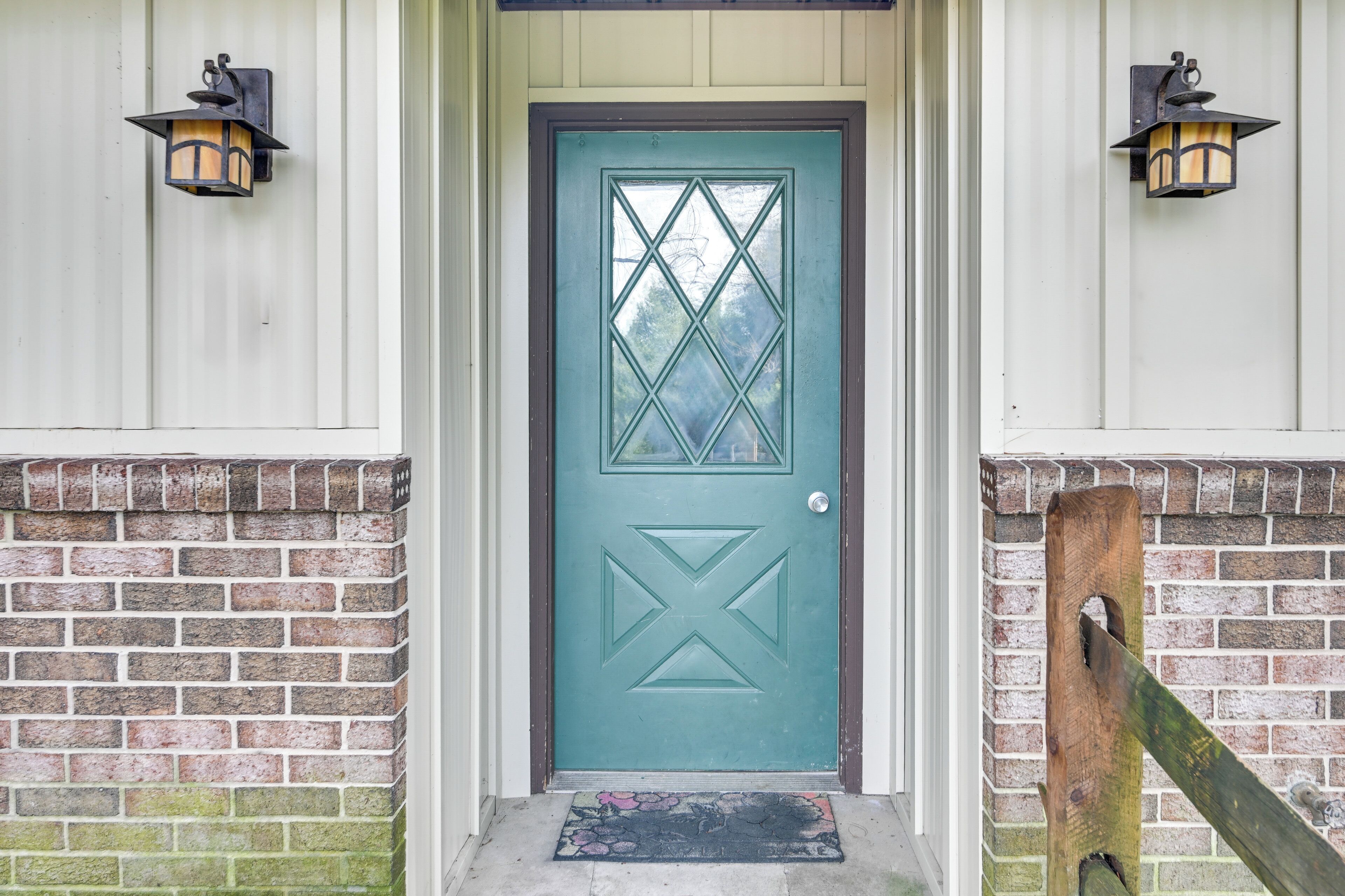 A teal door with a diamond pattern, flanked by two wall-mounted lanterns, leading to a brick wall entrance.