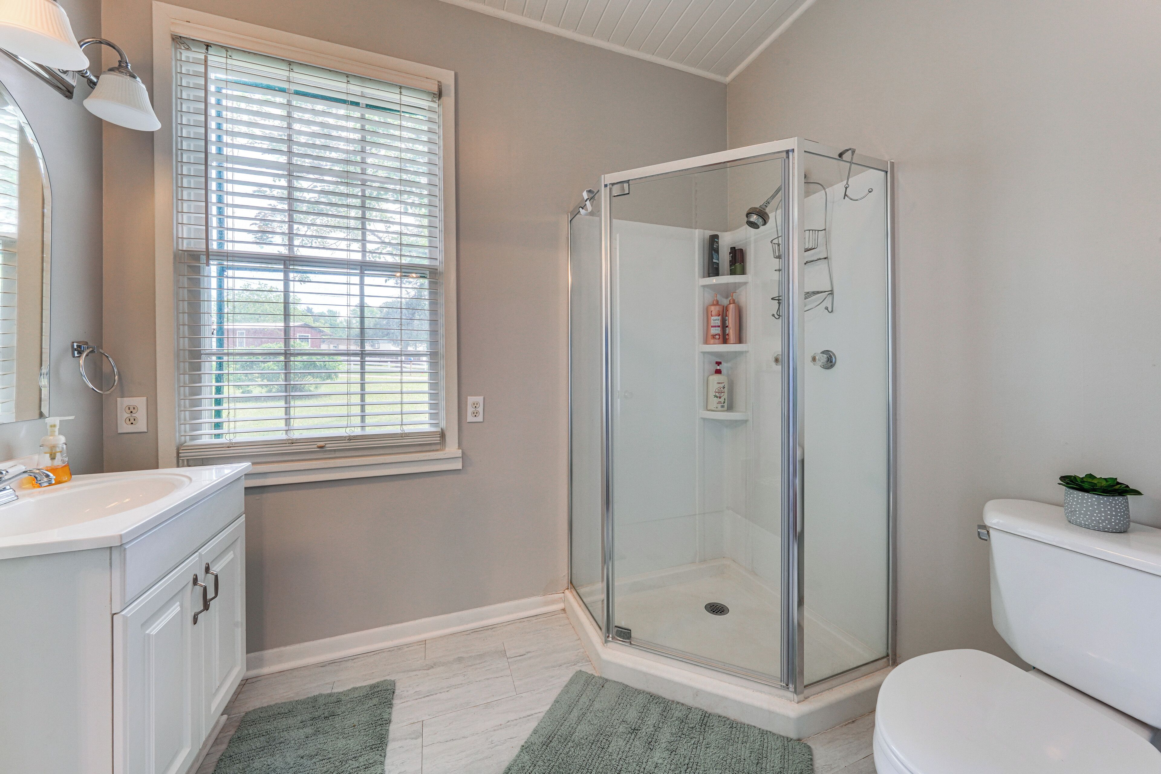 A bathroom with a glass shower enclosure, a toilet, a sink with a mirror, and a window with blinds.