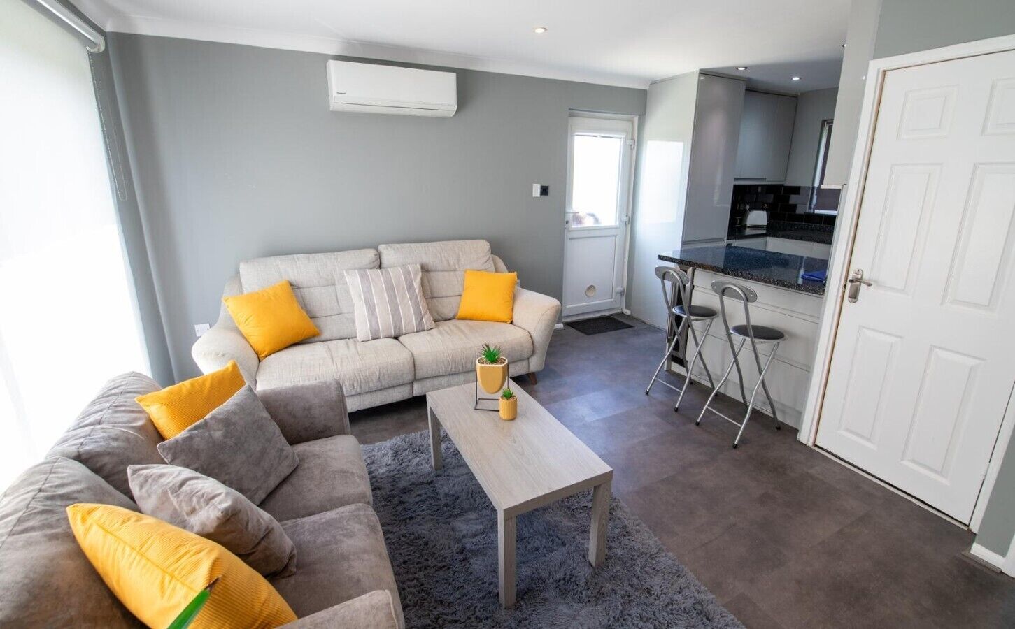 A modern living room with a grey sofa, yellow cushions, a coffee table with a plant, and a kitchen area with a bar stool.