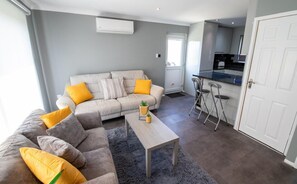 A modern living room with a grey sofa, yellow cushions, a coffee table with a plant, and a kitchen area with a bar stool. - Yew 2 Bungalow, Parkdean Resorts - Camber Sands (Camber)