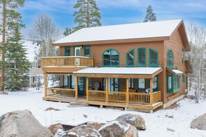 Una cabaña de madera de dos pisos con balcón, ventanas grandes y techo cubierto de nieve.