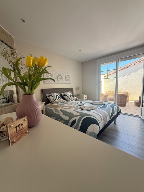 A bedroom with a bed, a nightstand, a lamp, a vase with flowers, and a view of the outside.