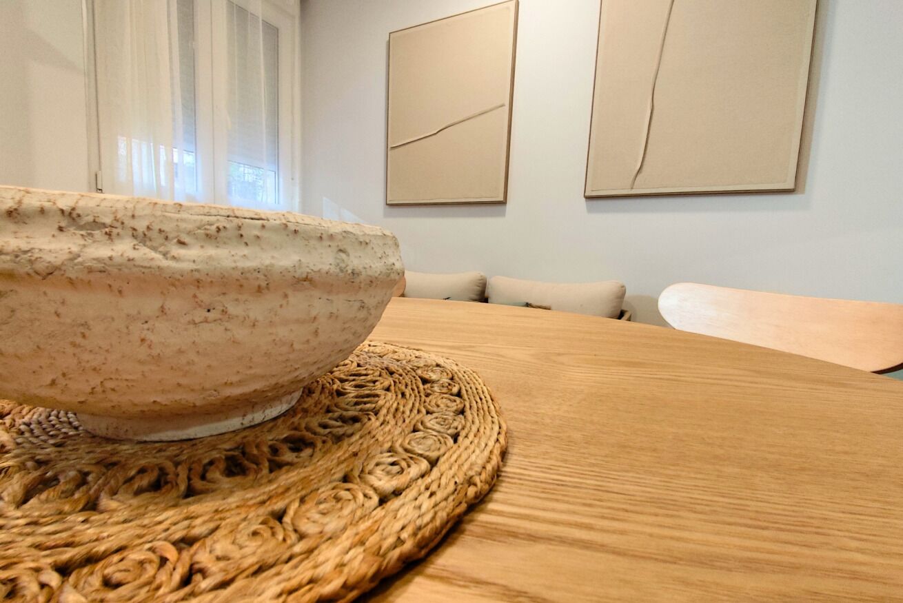 A wooden table with a woven placemat, a ceramic bowl, and a light-colored sofa in a room with light walls and abstract paintings.