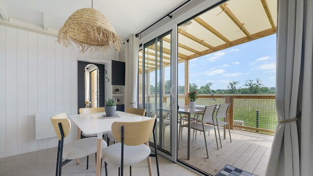Uma área de refeições com mesa e cadeiras, um lustre e vista para um deck externo através de uma porta de vidro.