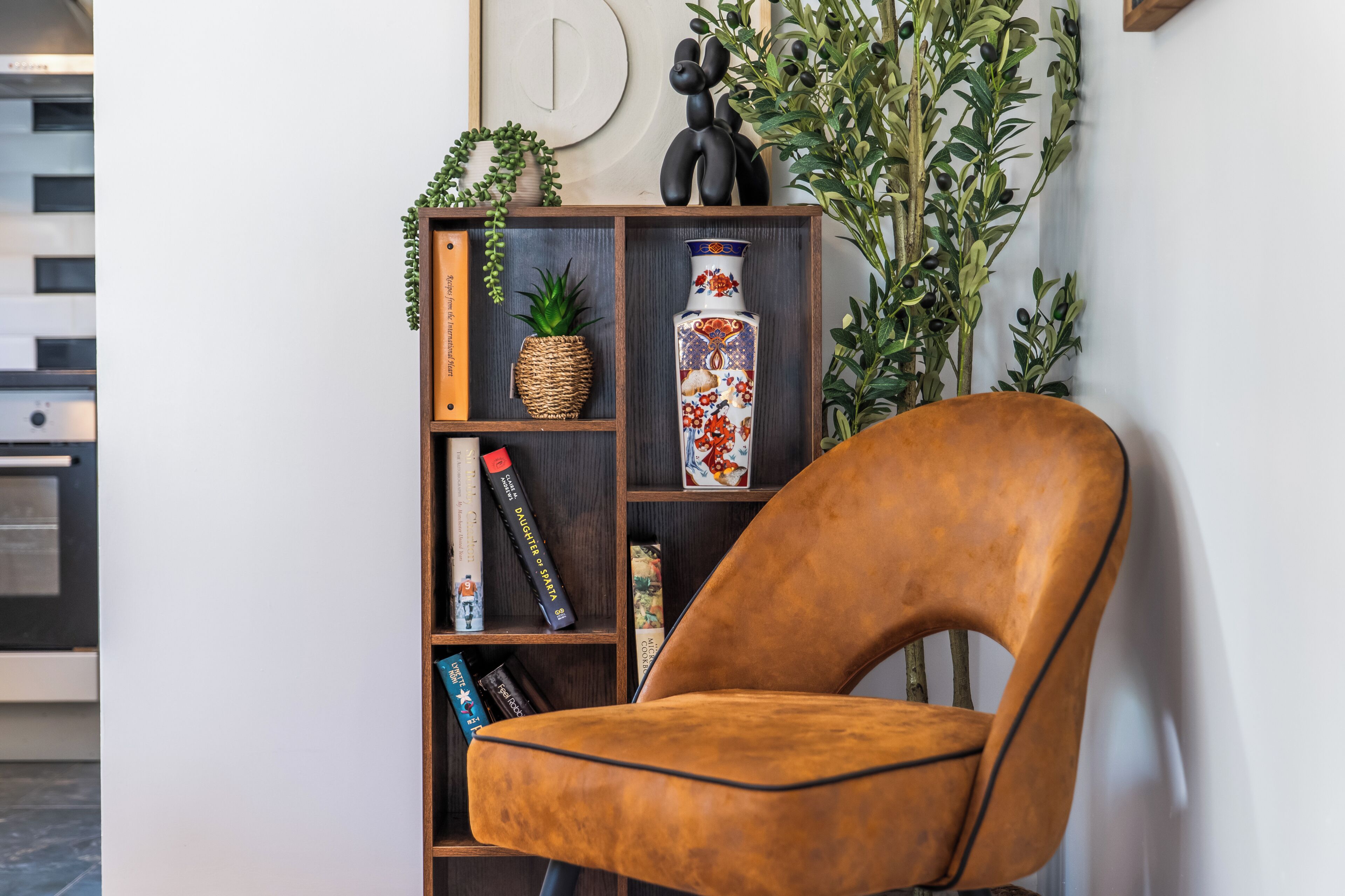 Een modern interieur met een bruine leren fauteuil, een houten boekenkast met decoratieve objecten, en een wandbevestigde letter 'D'.
