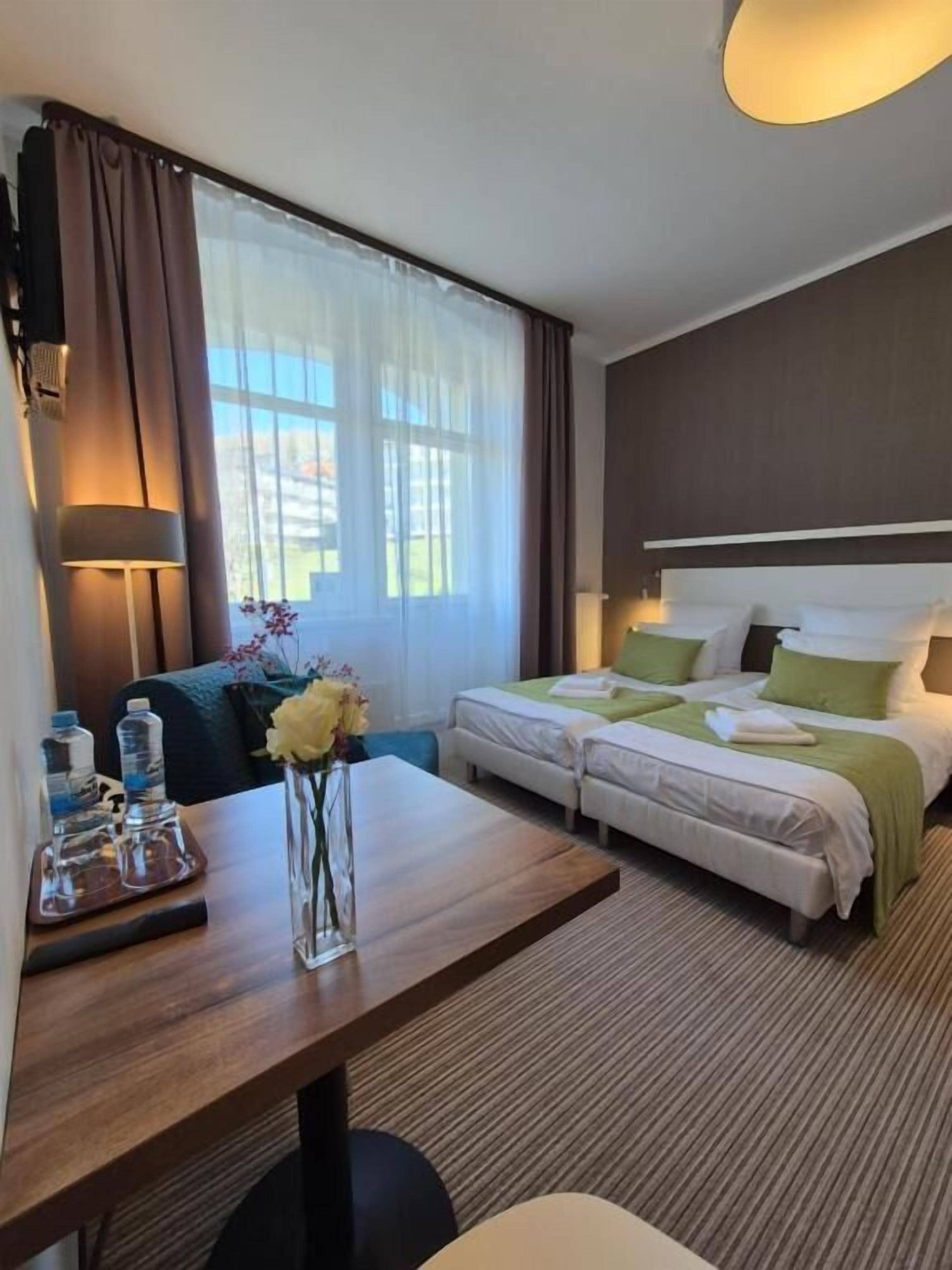 Ein Hotelzimmer mit einem Bett, einem Schreibtisch, einem Stuhl, einer Vase mit Blumen und einem fenster mit Vorhängen.