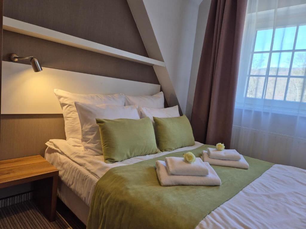 Hotellihuone, jossa on sänky, tyynyjä, yöpöytä, lamppu ja ikkuna, jossa on verhot.