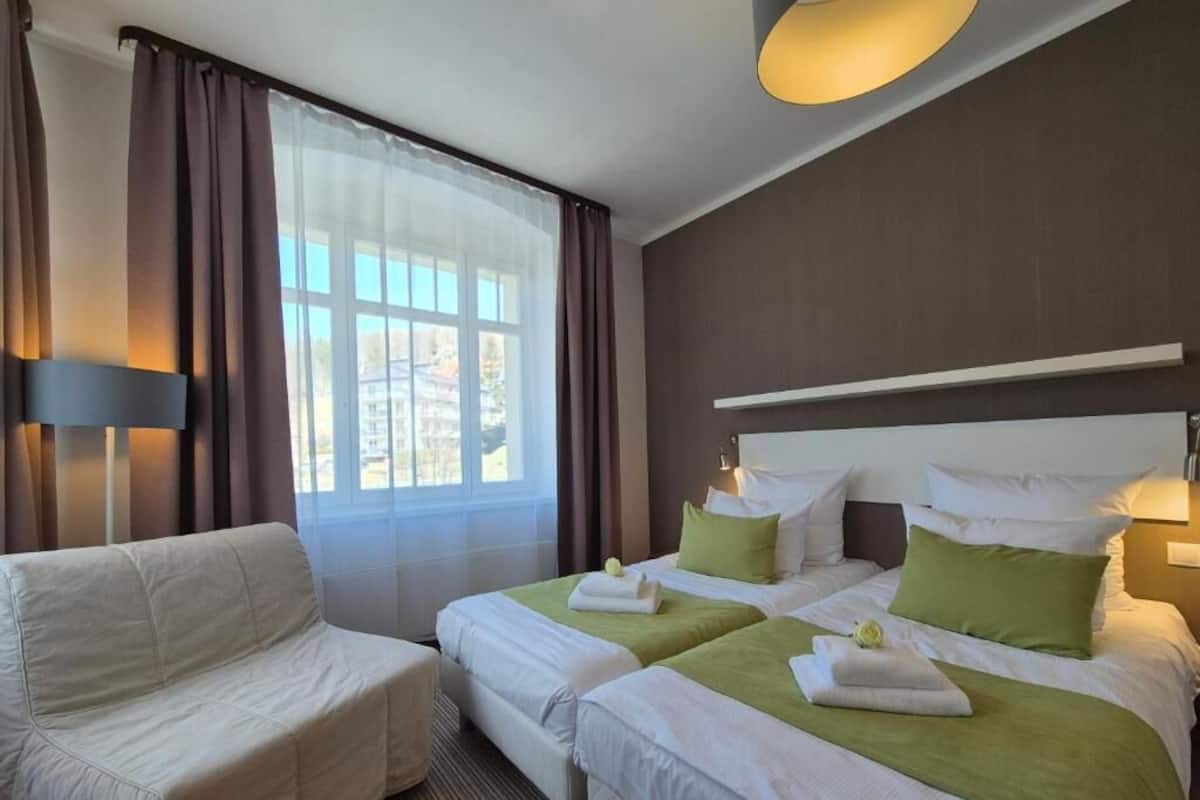 Quarto de hotel com duas camas, um sofá, uma janela grande e um abajur.