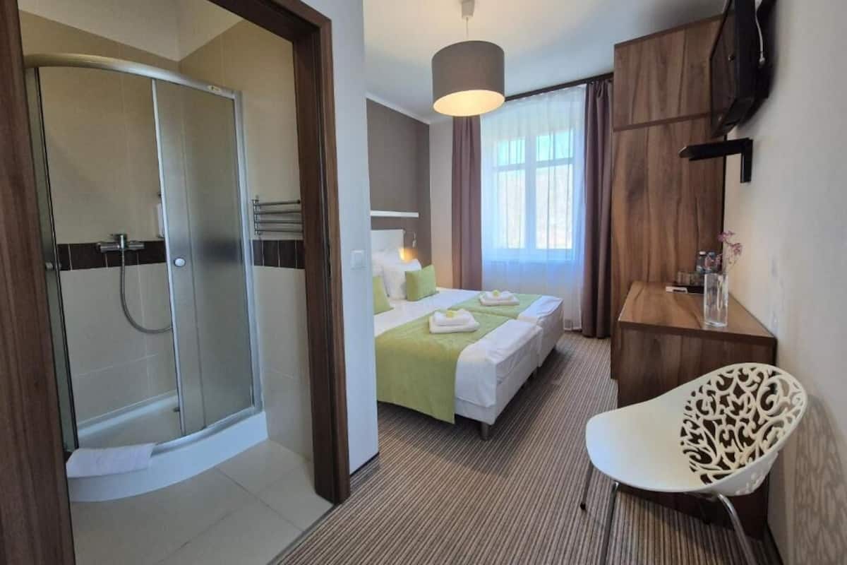 Quarto de hotel com chuveiro, uma cama com roupa de cama verde, escrivaninha com cadeira e televisão na parede.