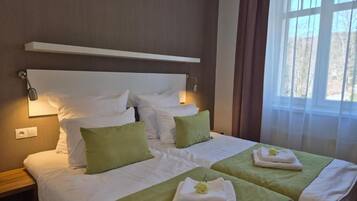 Une chambre d’hôtel avec un grand lit, deux tables de chevet, une petite table, une fenêtre avec des rideaux et une étagère fixée au mur.
