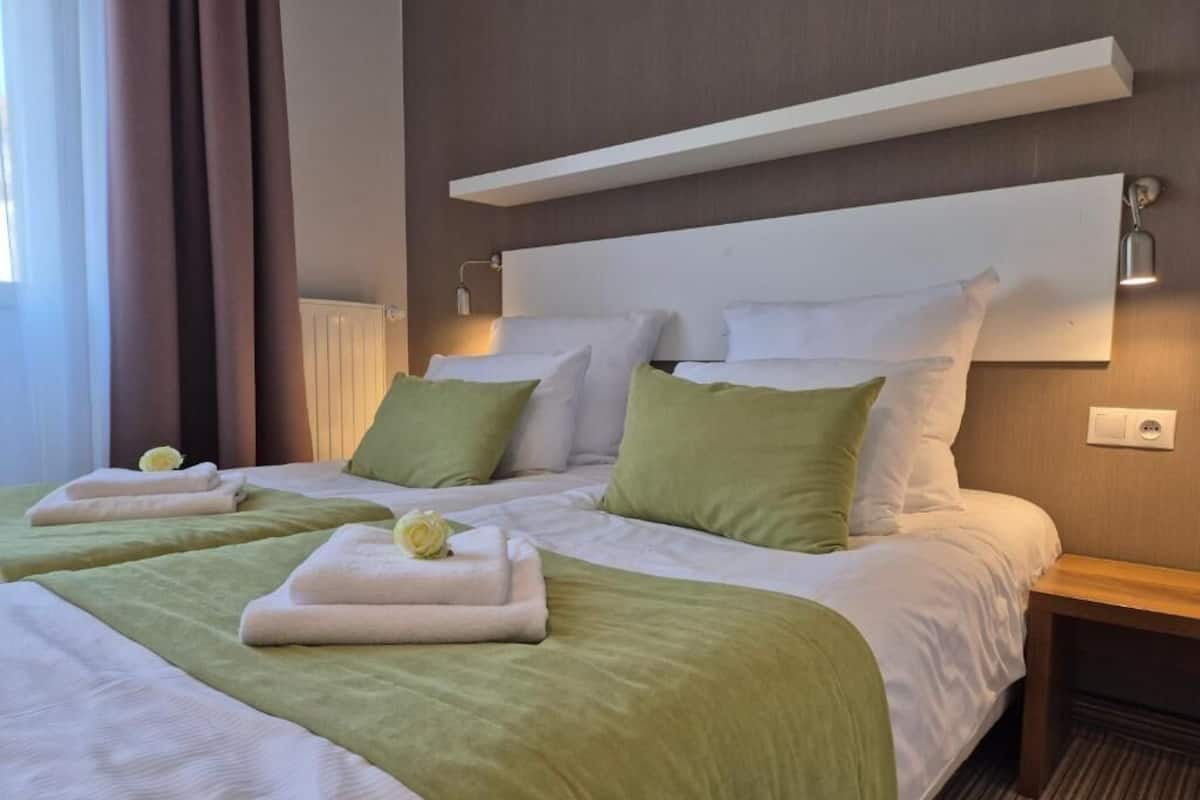 Quarto de hotel com duas camas, roupa de cama verde e branca, uma mesinha de cabeceira e um criado-mudo.