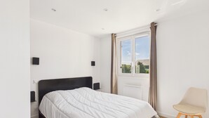 Un dormitorio con cama, una silla, un ventanal con cortinas y vistas al verdor exterior.