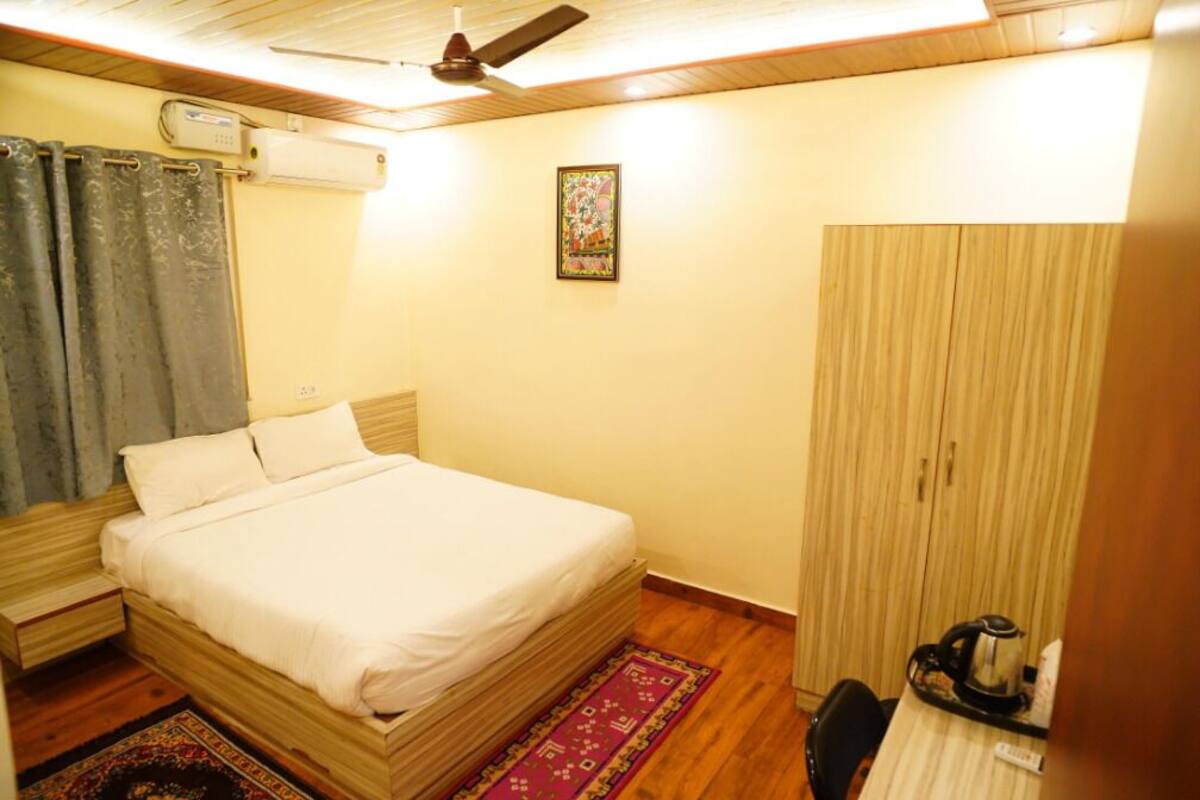 A single bed with a headboard, a ceiling fan, a wall-mounted air conditioner, a wooden wardrobe, and a small table with a kettle.