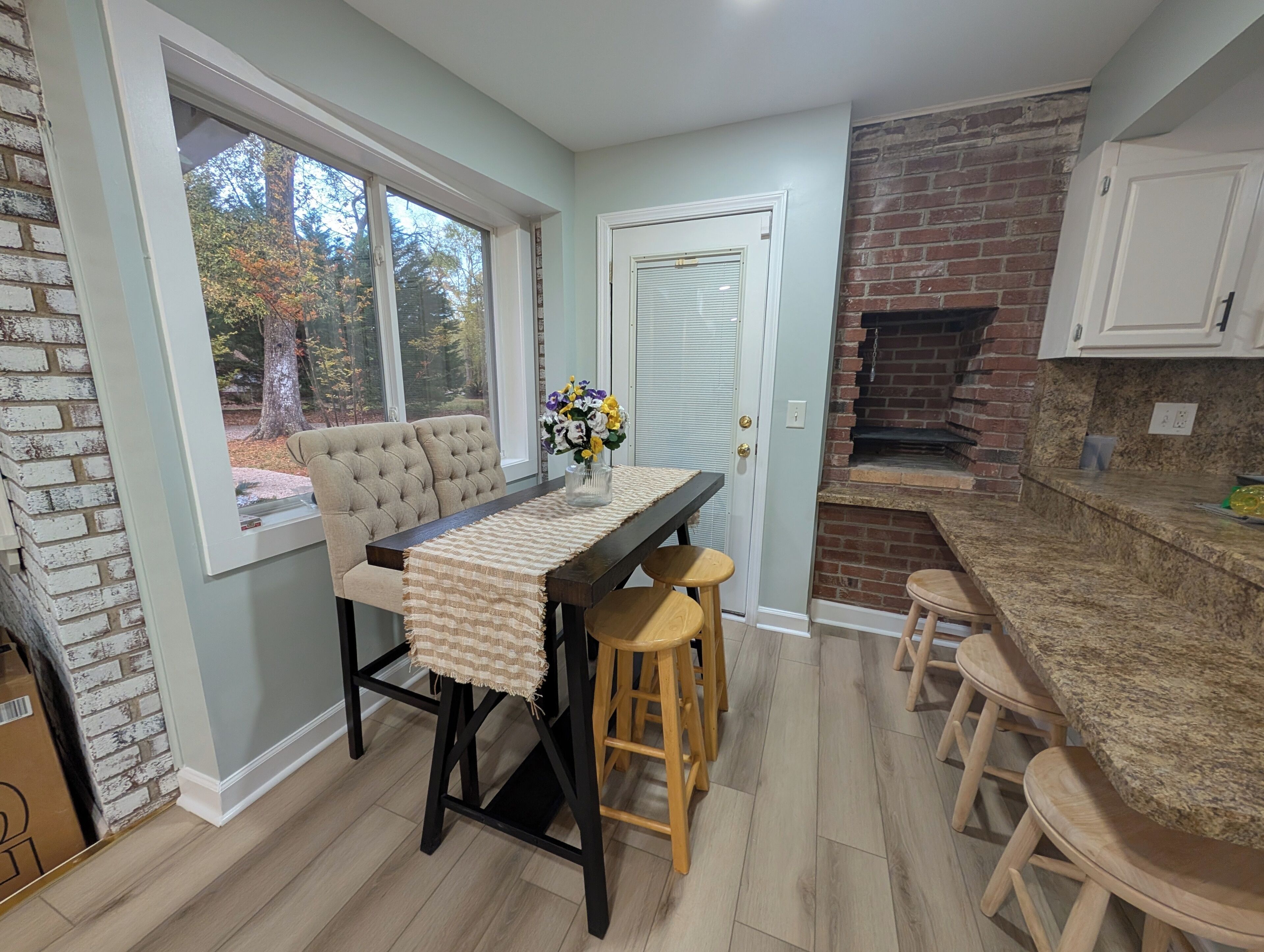A kitchen with a brick wall, a dining table with stools, and a view of trees through a window.