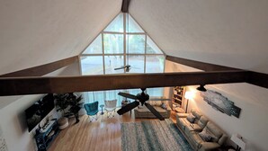 Una habitación estilo loft con techo inclinado, un ventanal grande, un ventilador de techo y una zona de estar con sofá y sillones.