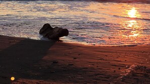En strand med vågor, en klippa och solen som går ner över havet.