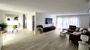 Uma sala de estar moderna com sofá, mesa de centro e televisão. Há uma secretária com cadeira e uma planta. O quarto tem soalho de madeira e paredes brancas.