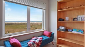 Ein Zimmer mit blauem Sofa, einem Fenster mit Jalousien, einem Holzregal und Blick auf den Strand.
