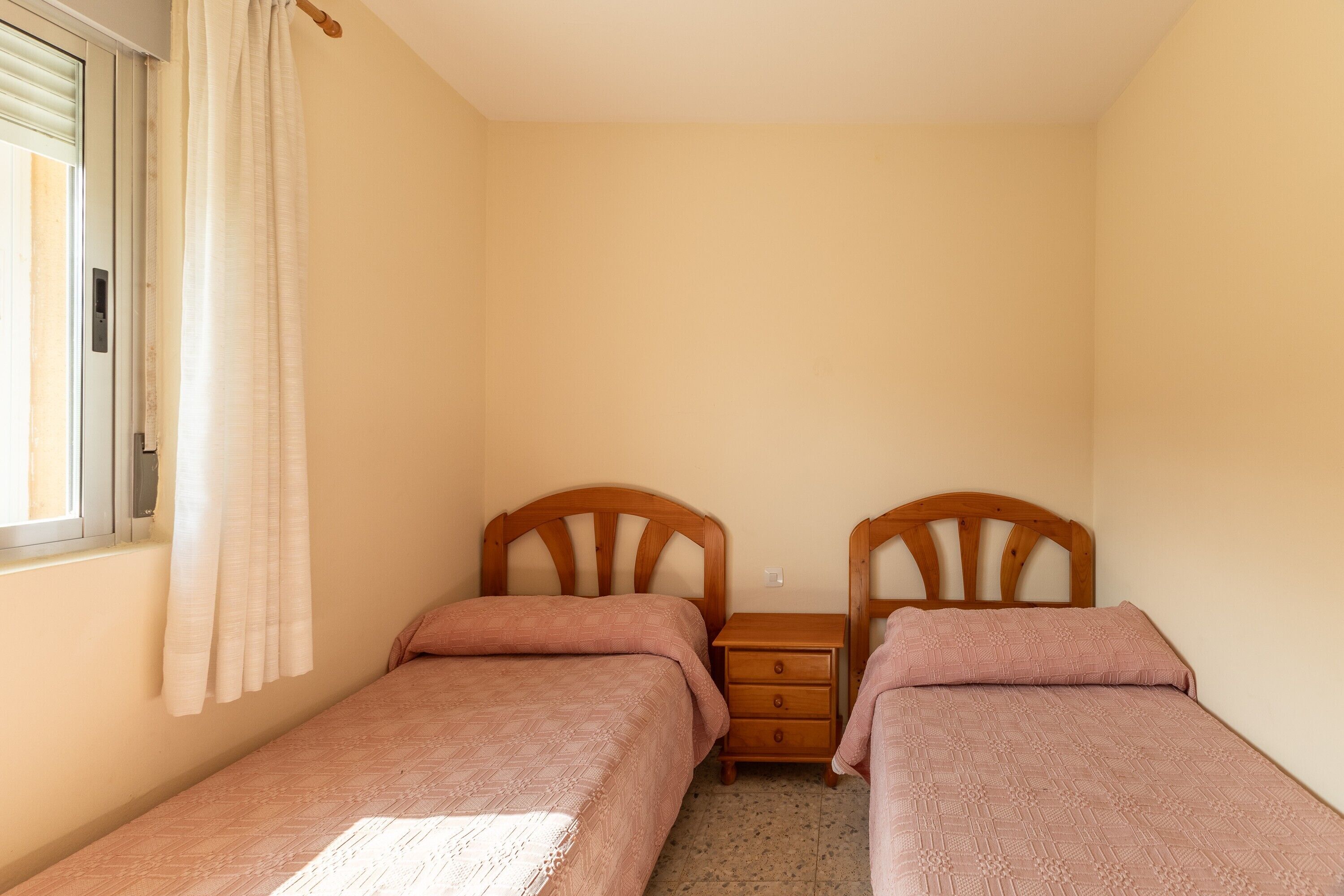 Un dormitorio con dos camas individuales, una mesita de noche de madera, una ventana con cortinas y piso de baldosas.
