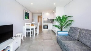 Una sala de estar moderna con un televisor de pantalla plana, un sofá gris, un comedor con sillas blancas, una cocina con armarios blancos y una planta en maceta.
