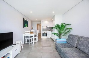 Un salon moderne avec un téléviseur à écran plat, un canapé gris, un coin repas avec des chaises blanches, une cuisine avec des meubles blancs et une plante en pot.