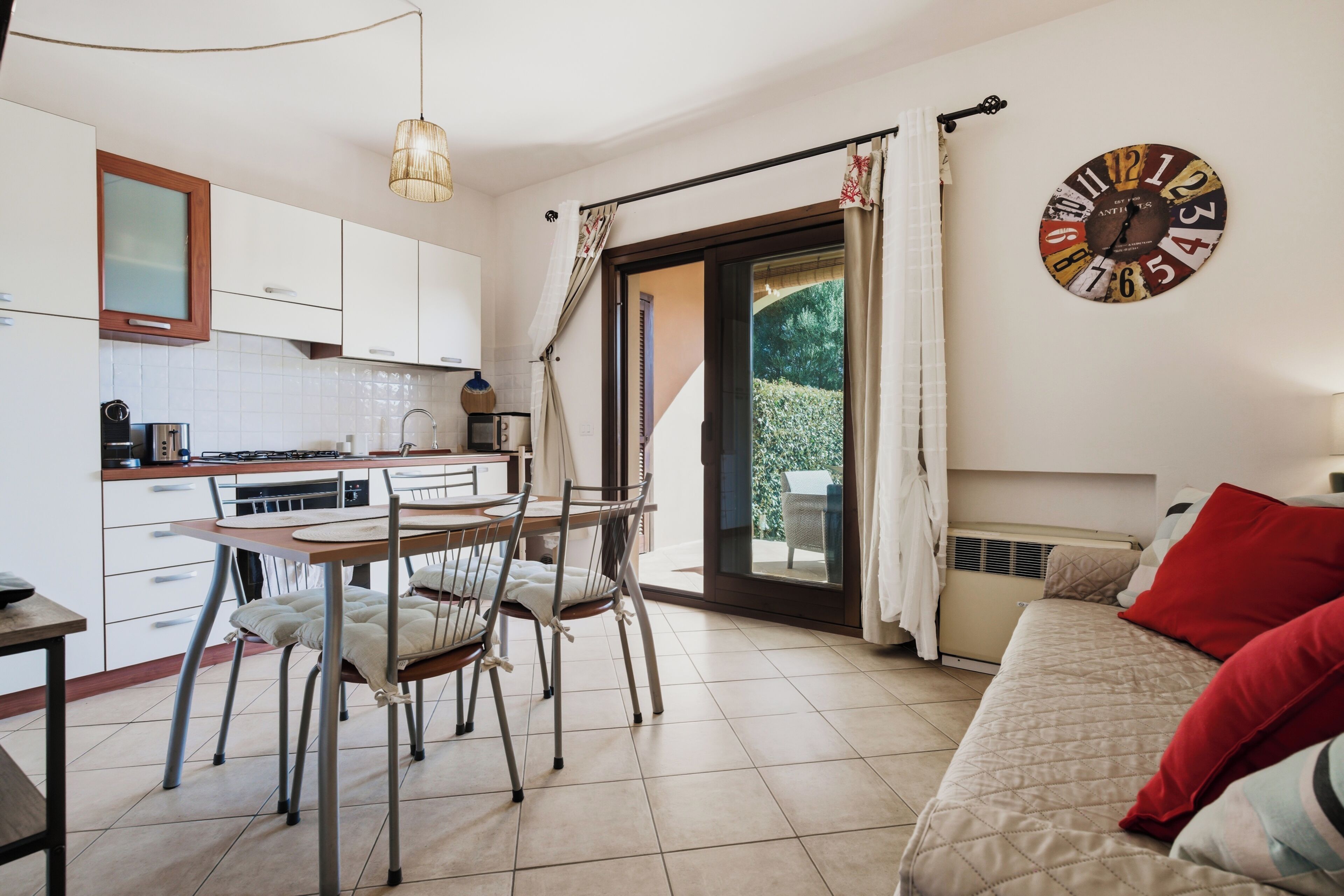 Een compacte keuken met moderne apparaten, een zithoek met een tafel en stoelen, een bed met rode kussens en een grote wandklok.