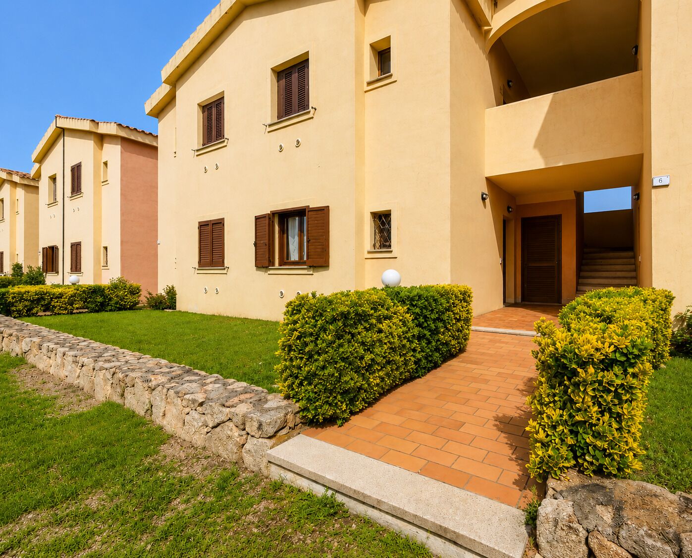 A multi-story building with a stone pathway leading to the entrance, surrounded by well-maintained hedges and a stone wall.