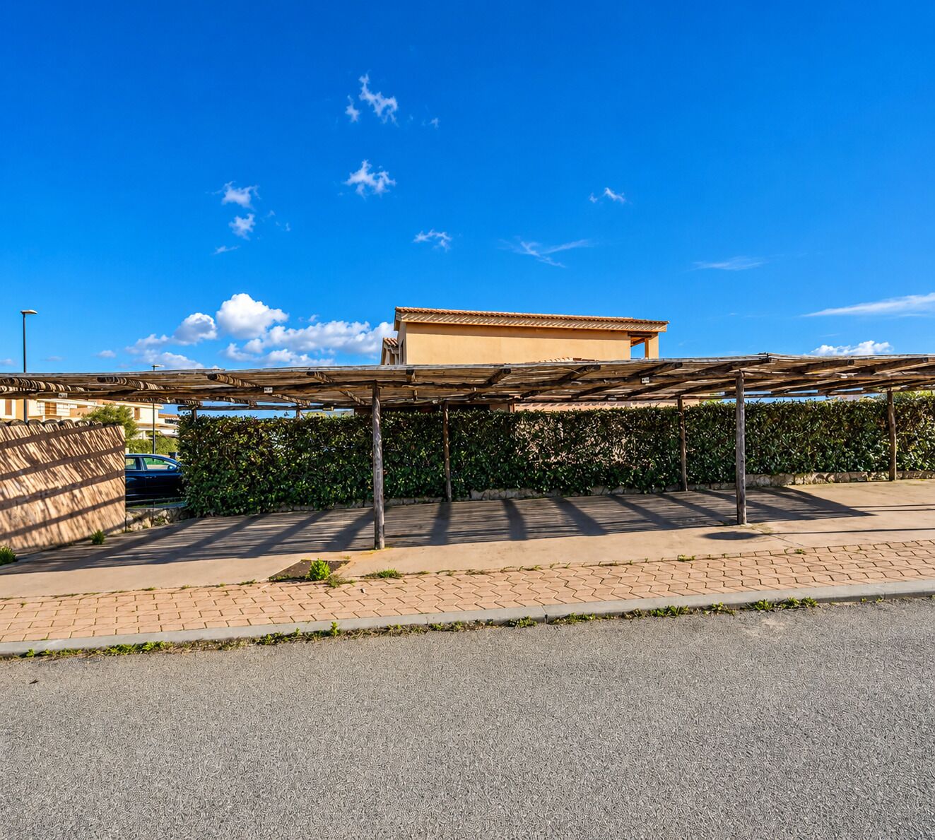 A covered outdoor area with a pergola structure, a paved walkway, and a building with a tiled roof in the background.
