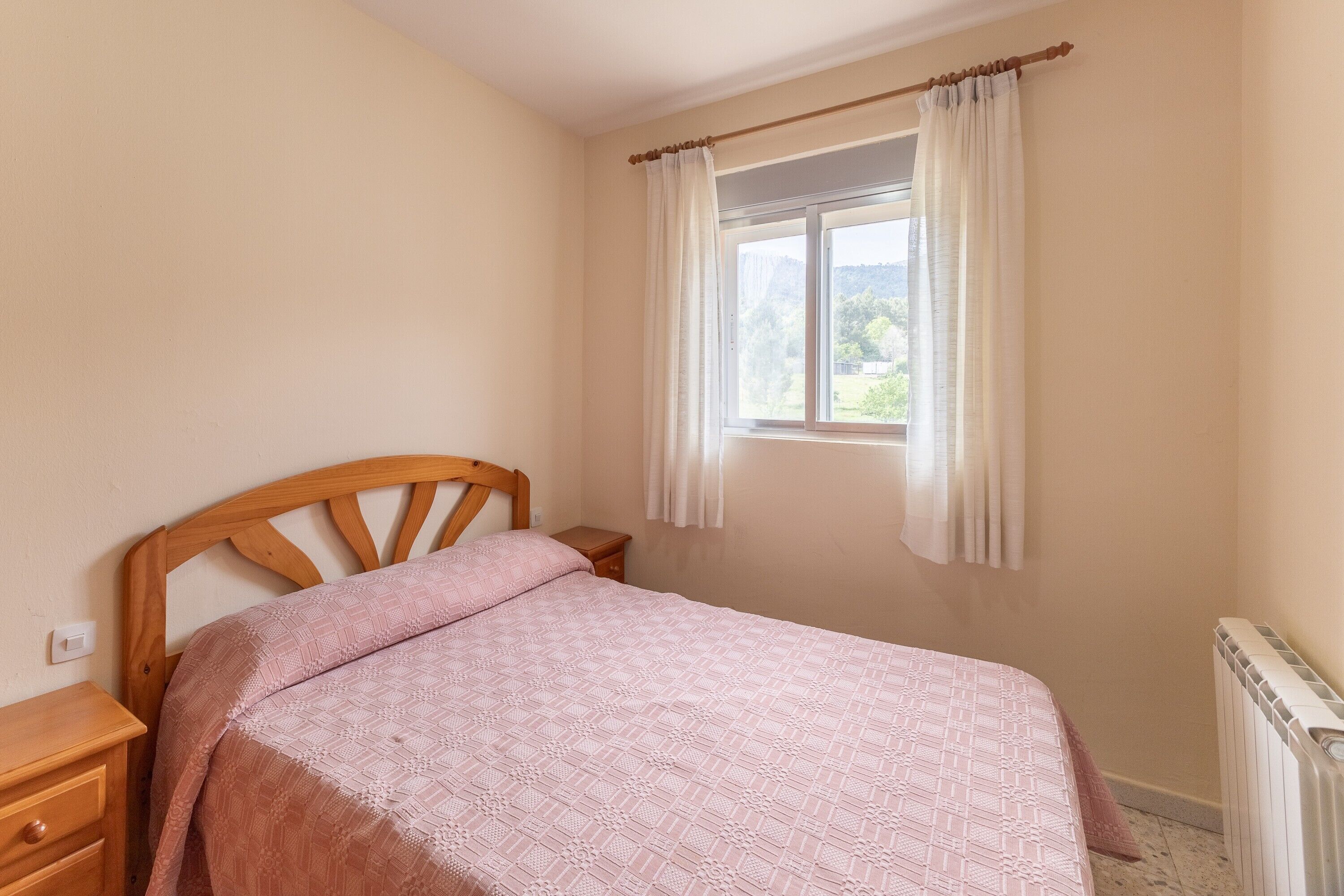 Un dormitorio con una cama de madera, una mesita de noche, un ventanal con cortinas y un radiador.