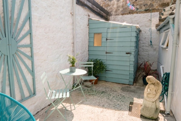 Un pequeño patio al aire libre con una mesa y sillas blancas, un cobertizo azul y una estatua.