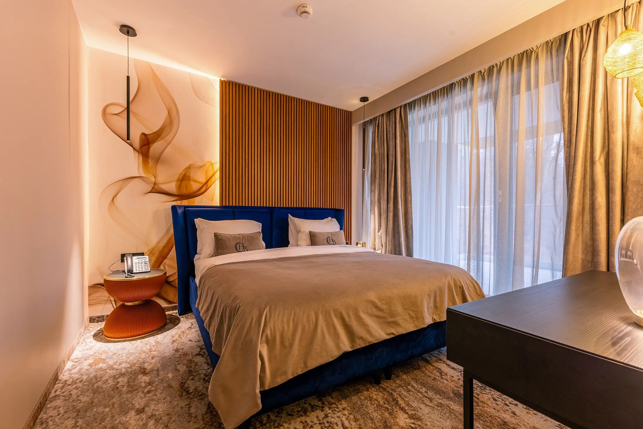 A modern hotel room with a large bed, a side table with a phone, a desk, and a wall-mounted light fixture.