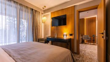 A hotel room with a large bed, a desk with a lamp, a TV, and a view of the outdoors through sheer curtains.