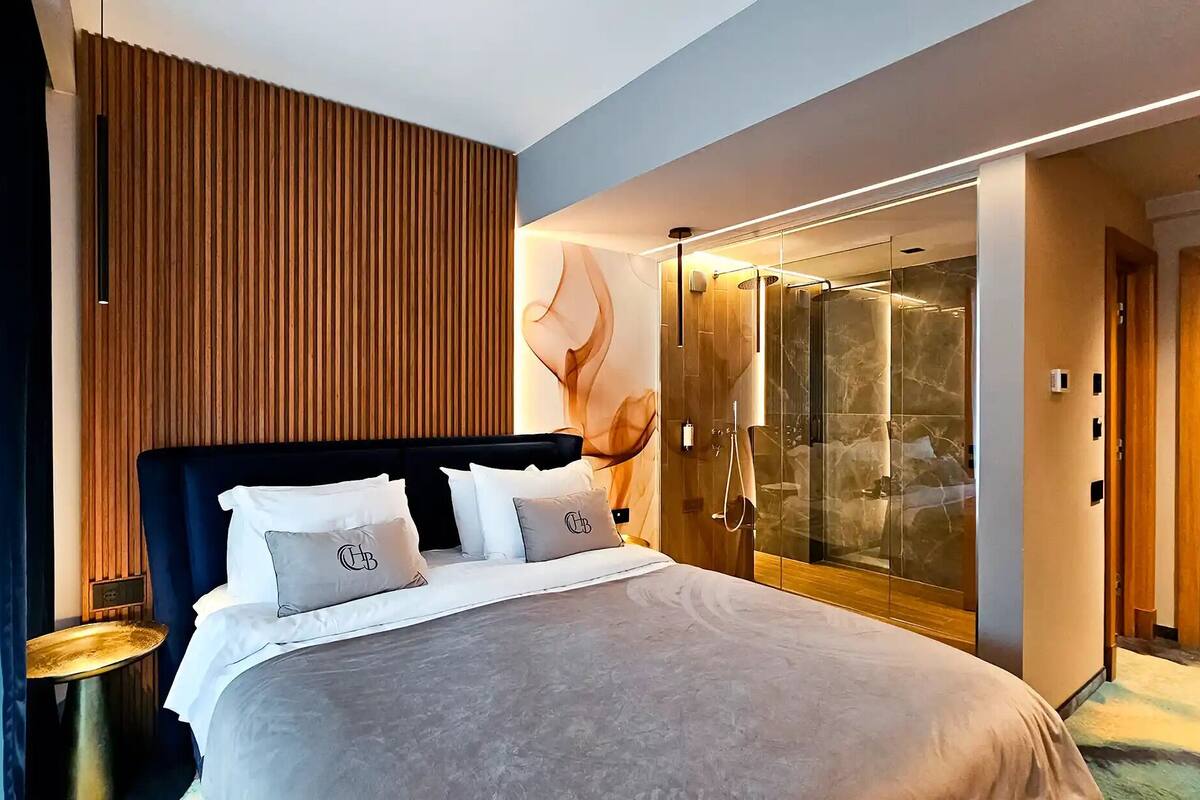 Una habitación de hotel moderna con una cama grande, un vestidor y una ducha con mampara de vidrio.