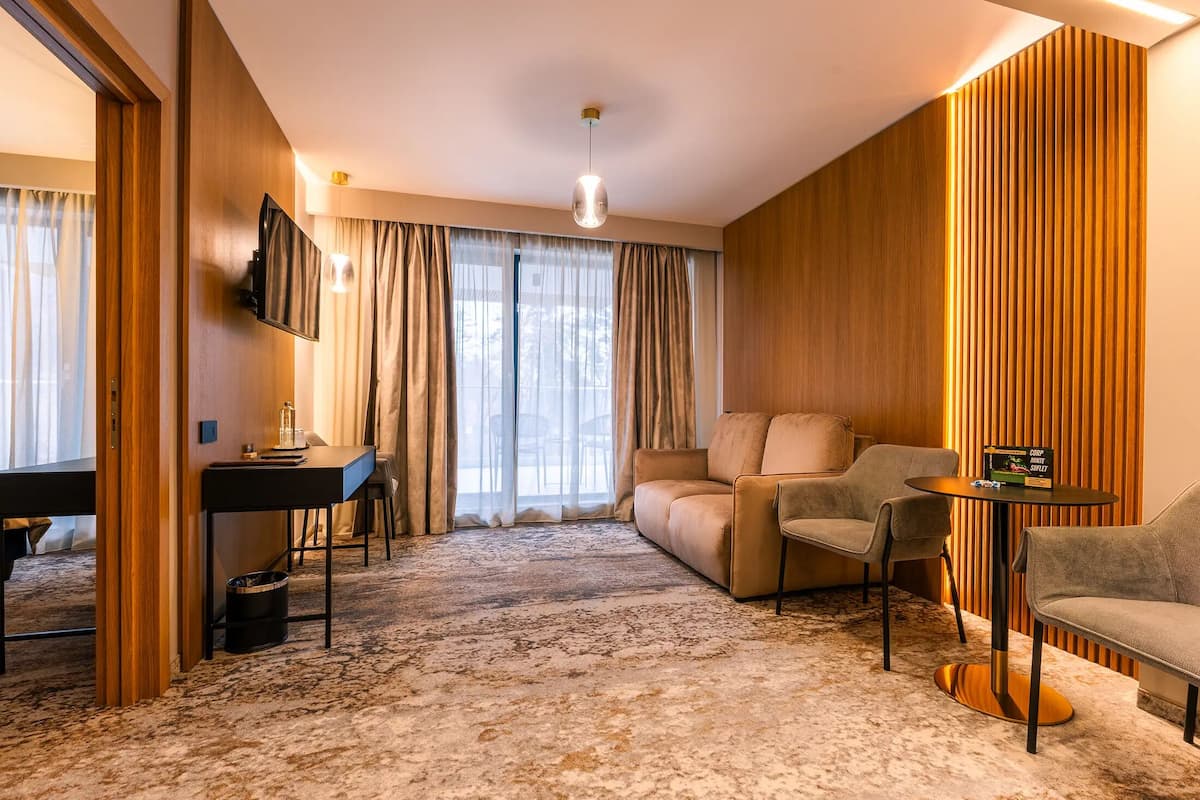 Habitación de hotel con escritorio, sofá, sillas, televisor y una ventana grande con cortinas.
