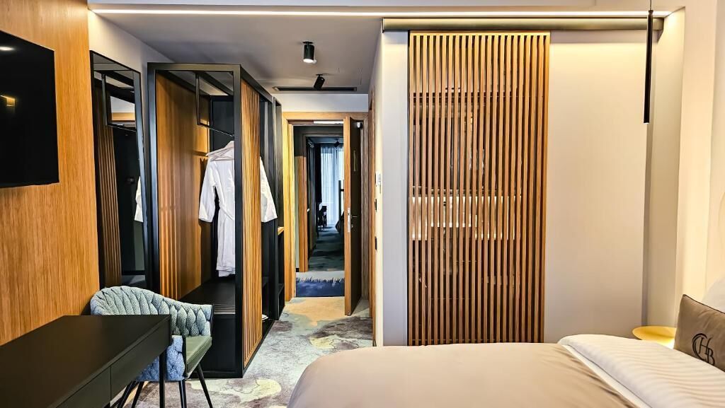 A modern hotel room with a bed, a desk, a chair, a TV, and a wardrobe.