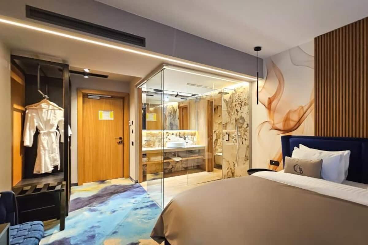 Habitación de hotel con cama, perchero para bata, baño con paredes de vidrio y una pared con paneles de madera.