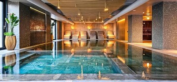 Una amplia piscina interior con superficie de agua cristalina, rodeada de zonas de descanso y paredes con texturas decorativas.