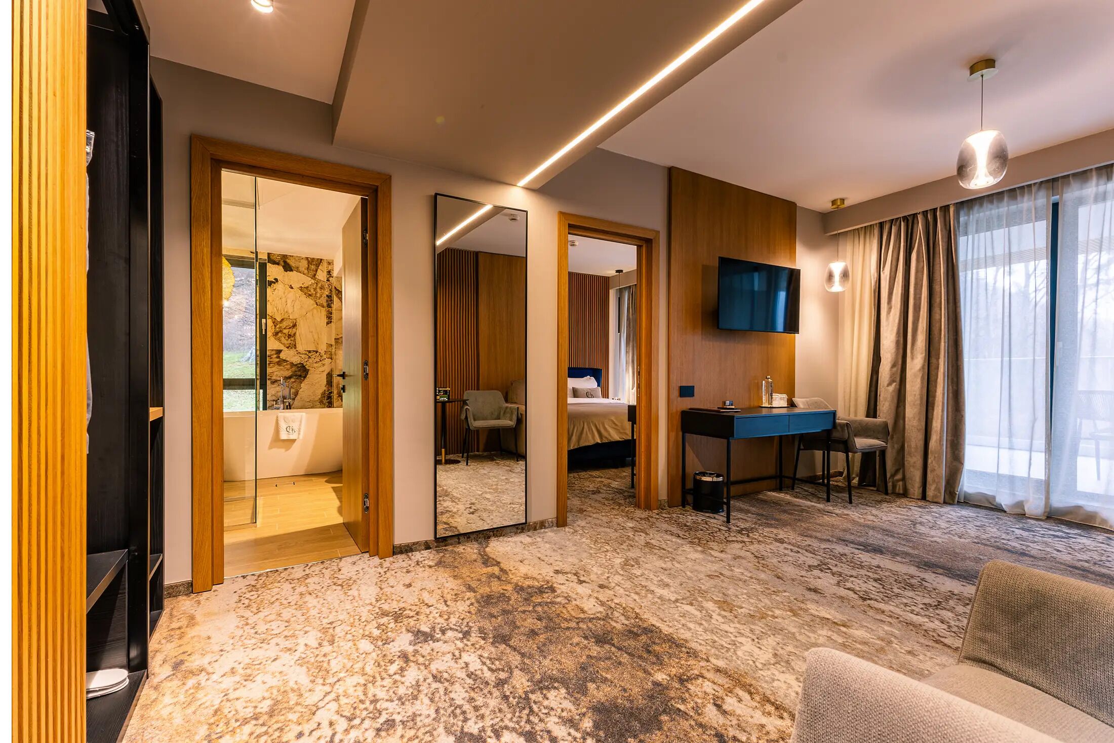 A hotel room with a marble floor, a sofa, a desk with a chair, a TV, and a bathroom visible through a glass door.