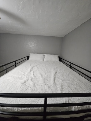 A single bed with a white quilt, black metal headboard and footboard, and a plain wall with a textured ceiling.