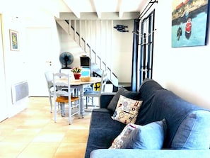 Una sala de estar con un sofá azul oscuro, una mesa de comedor con sillas, una escalera y un ventilador montado en la pared.