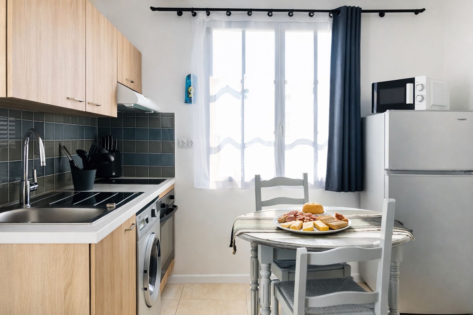 Cocina compacta con lavabo, estufa y microondas, una mesa de comedor con un plato de comida y una ventana con cortinas.
