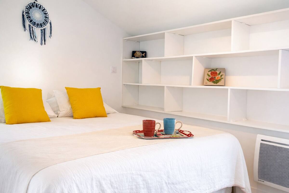 A hotel room with a bed, white walls, a decorative wall hanging, and a shelf with a few items.