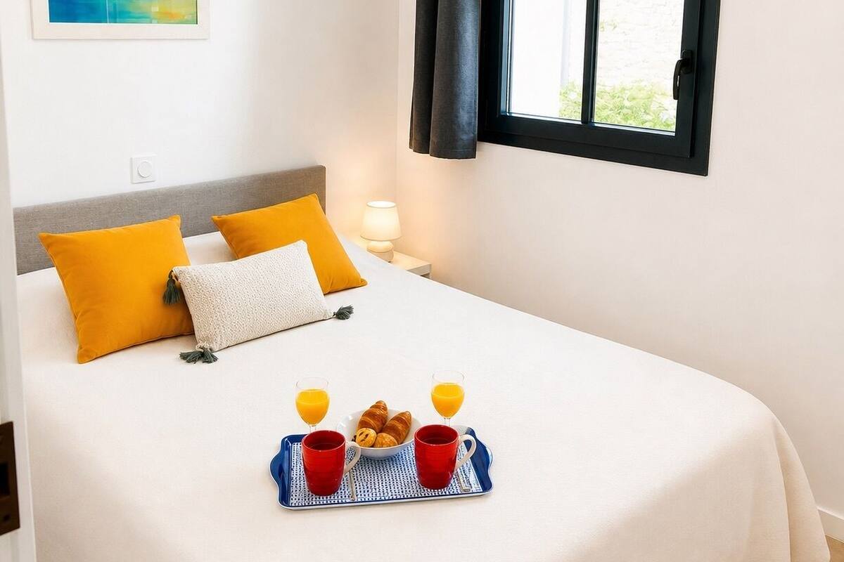 A neatly made bed with a tray of breakfast items, a painting on the wall, and a window with curtains.