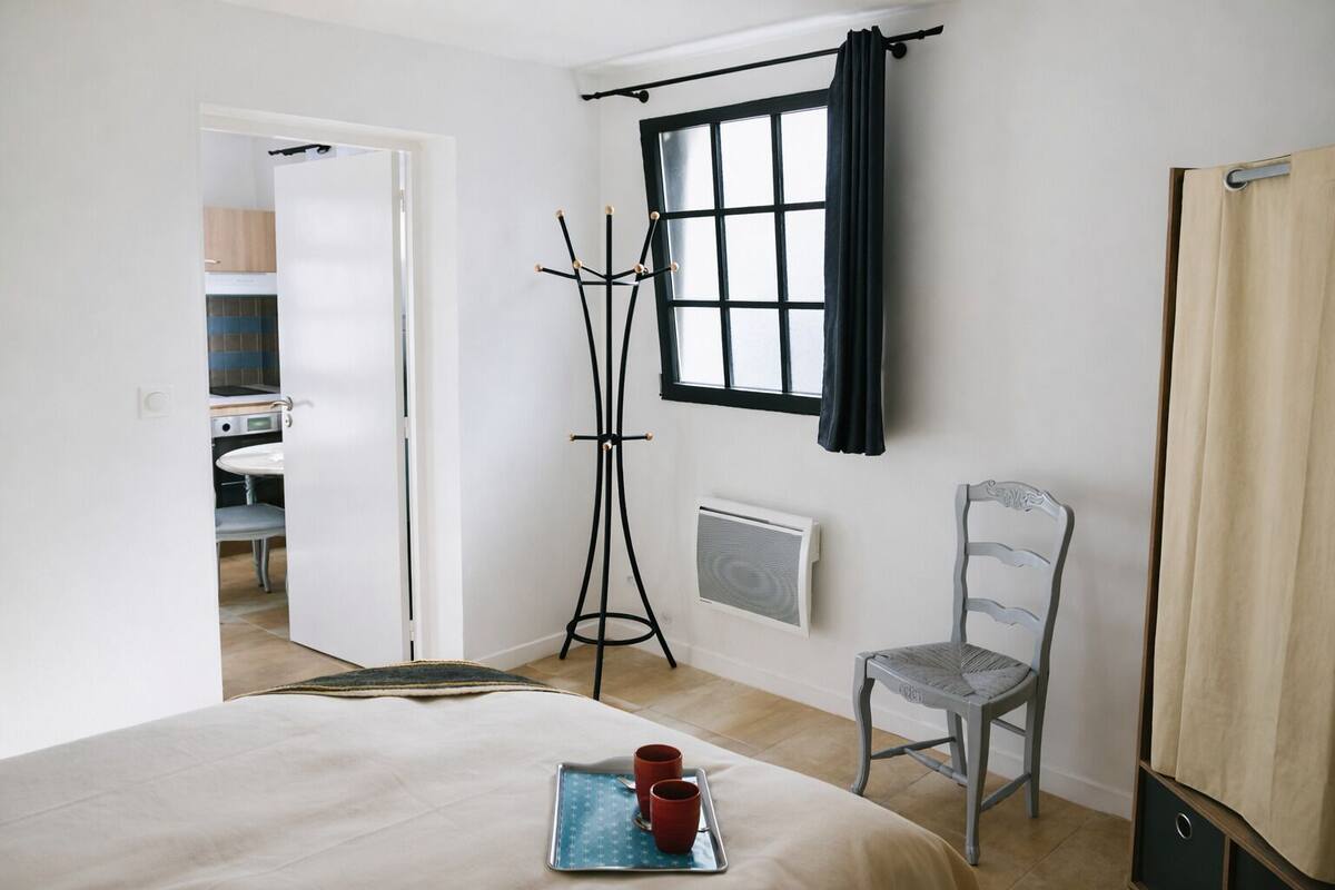 A bedroom with a bed, a chair, a coat rack, a window with curtains, and a radiator.