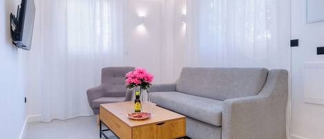A modern living room with a grey sofa, a wooden coffee table with a bottle and glasses, and a pink flower arrangement.